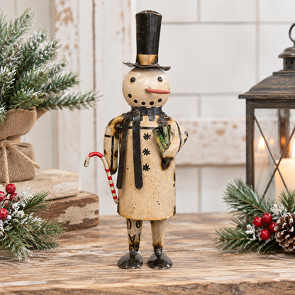 A decorative snowman figurine stands on a rustic wooden surface, wearing a tall black top hat, black-and-beige coat, and holding a red-and-white candy cane. The scene is styled with evergreen branches, red berries, stacked books, and a lantern in the background.