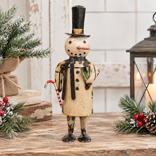 A decorative snowman figurine stands on a rustic wooden surface, wearing a tall black top hat, black-and-beige coat, and holding a red-and-white candy cane. The scene is styled with evergreen branches, red berries, stacked books, and a lantern in the background.