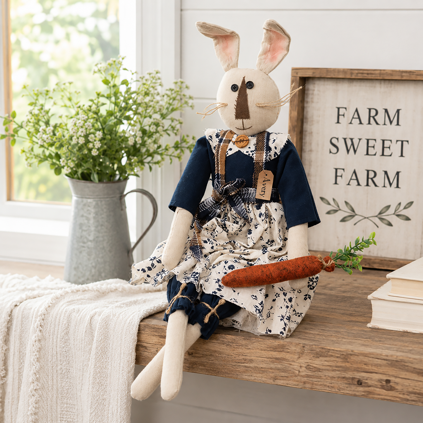 Avery bunny rag doll shelf sitter with ivory fabric, button eyes, posable ears, a navy blue dress with floral apron, plaid shoulder straps, and a bright orange felt carrot, creating a warm farmhouse, cottage, and spring home décor accent.
