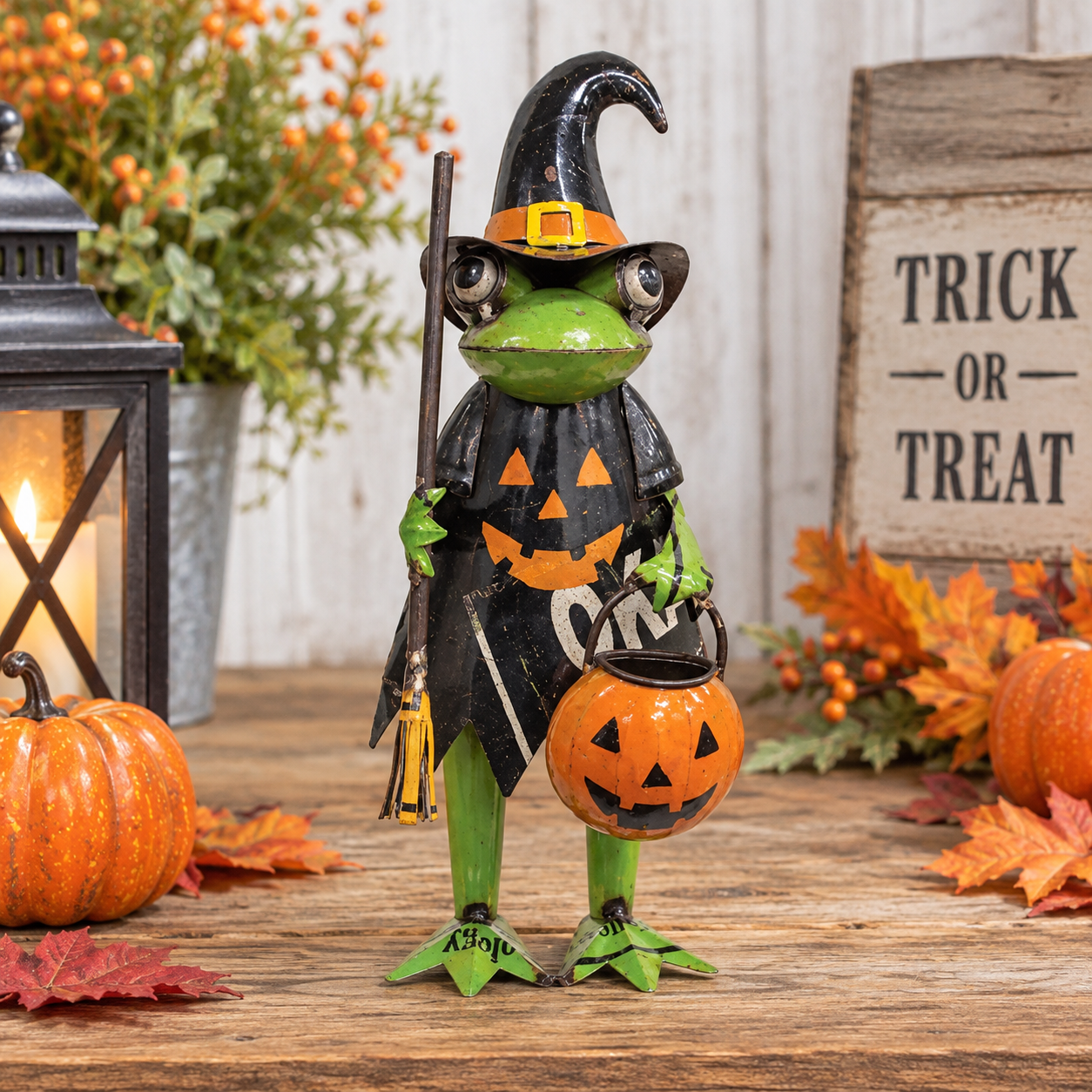 A Halloween frog figurine dressed as a witch, holding a jack-o'-lantern bucket, displayed on a rustic wooden surface with autumn decorations including pumpkins, leaves, a lantern, and a 'Trick or Treat' sign.