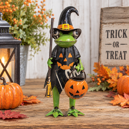 A Halloween frog figurine dressed as a witch, holding a jack-o'-lantern bucket, displayed on a rustic wooden surface with autumn decorations including pumpkins, leaves, a lantern, and a 'Trick or Treat' sign.