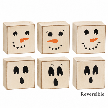 Six small square wooden blocks with painted faces, arranged in two rows on a white background. The top row shows snowman faces with black eyes, black dotted mouths, and orange carrot noses.