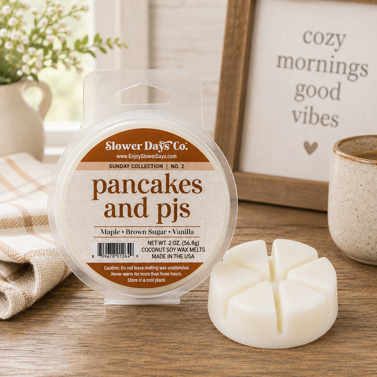 A round package of Slower Days Co. soy wax melts labeled 'pancakes and pj's' sits on a wooden table beside a white four-petal wax melt piece.