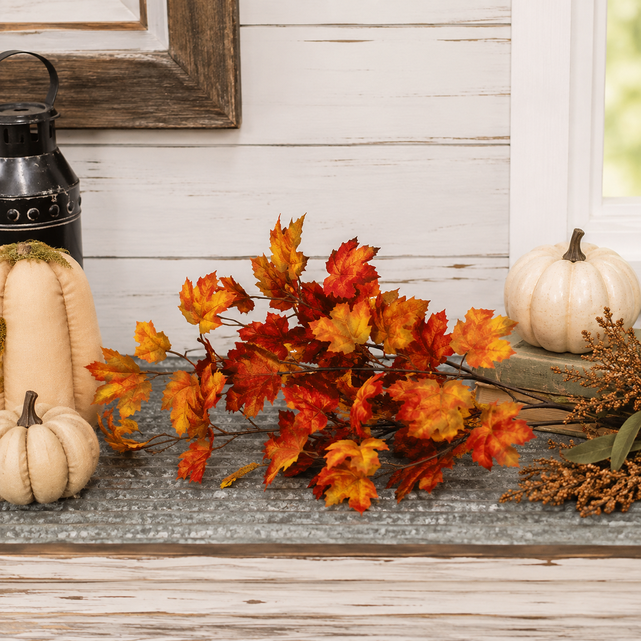 Autumn artificial leaf garland with vibrant orange, red, and yellow maple leaves arranged on a rustic gray tabletop, flanked by cream pumpkins and a black lantern against a white wood paneled wall.
