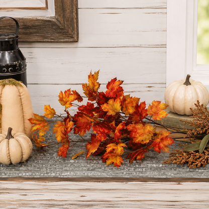 Autumn artificial leaf garland with vibrant orange, red, and yellow maple leaves arranged on a rustic gray tabletop, flanked by cream pumpkins and a black lantern against a white wood paneled wall.