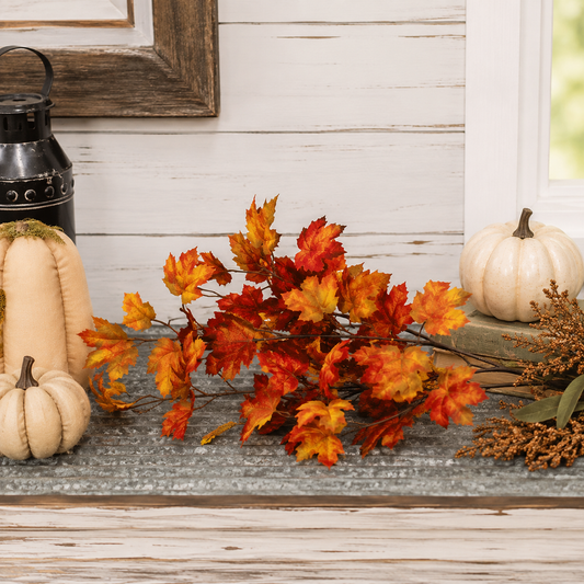 Autumn artificial leaf garland with vibrant orange, red, and yellow maple leaves arranged on a rustic gray tabletop, flanked by cream pumpkins and a black lantern against a white wood paneled wall.