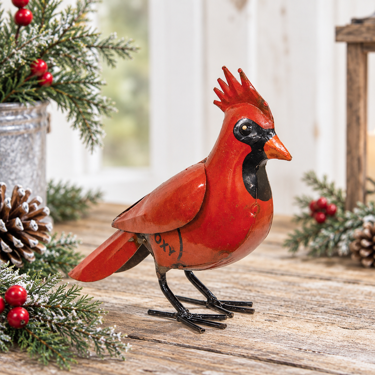 A decorative red cardinal bird figurine with black details, standing on a rustic wooden surface and surrounded by holiday greenery, pinecones, and red berries.