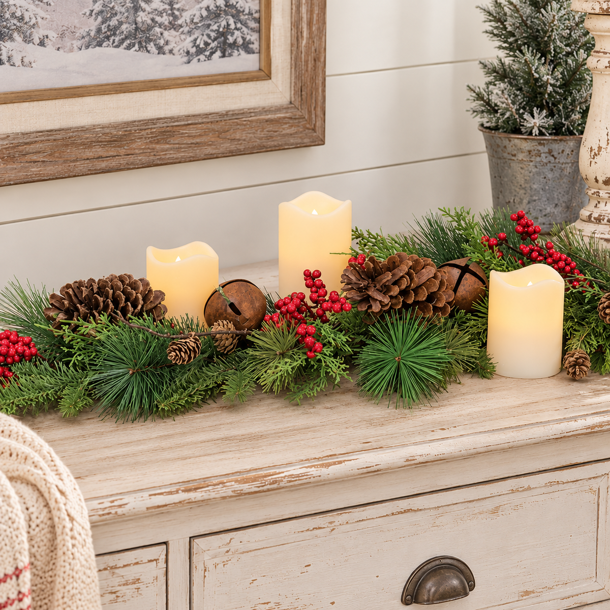A festive Christmas garland arranged on a distressed white wooden chest, featuring green pine branches, brown pinecones, small brown bells, red berry clusters, and three cream-colored flameless LED candles.