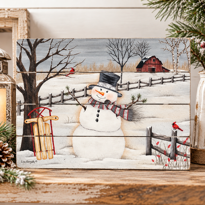 A square wooden sign with a winter landscape painting featuring a snowman, bare trees, a red barn, fence, sled, and small red birds, displayed on a rustic wooden surface with holiday decor.