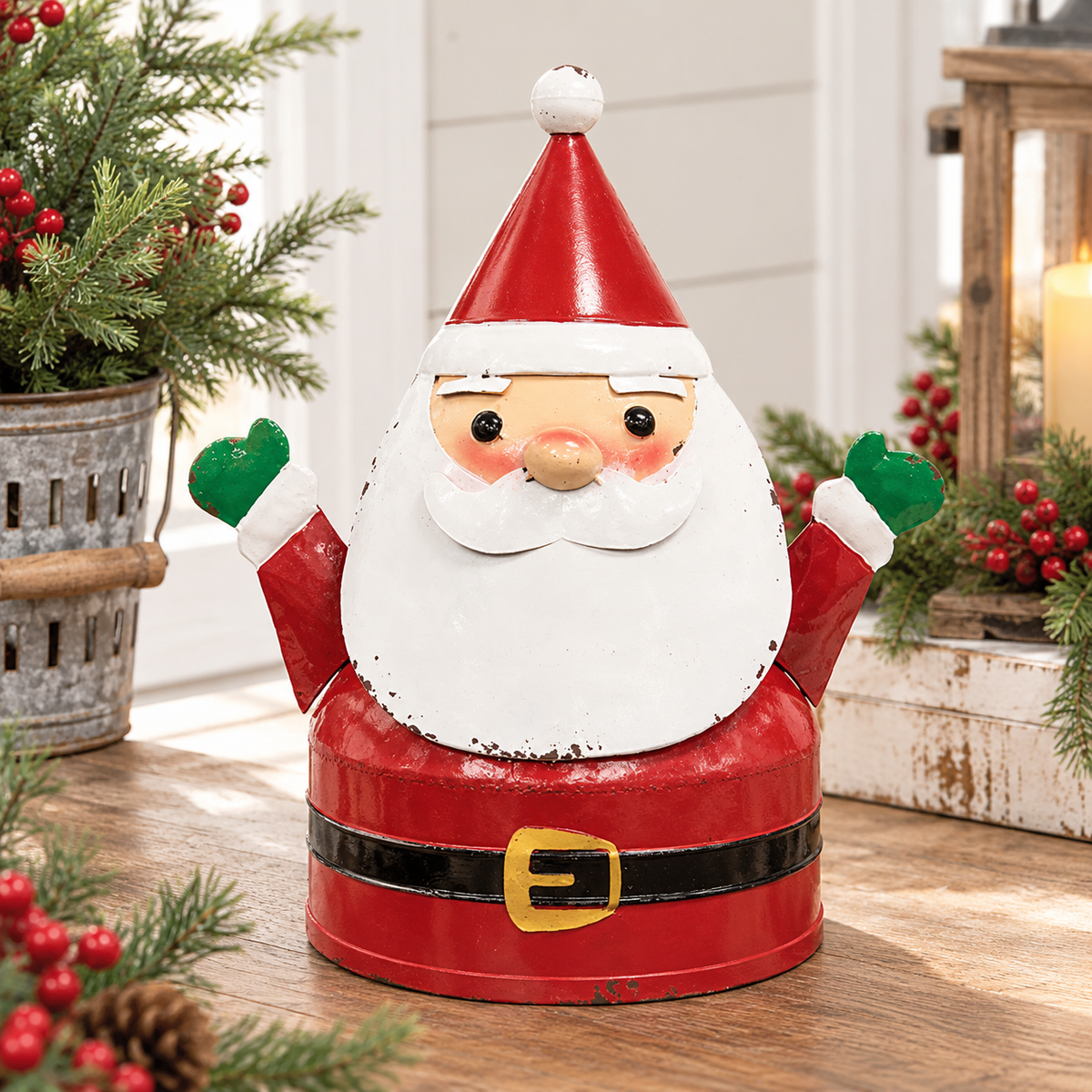 A festive metal Santa Claus ornament or container with a distressed red and white finish, green mittens, a black belt, and a gold buckle, displayed on a wooden table with holiday decorations in the background.