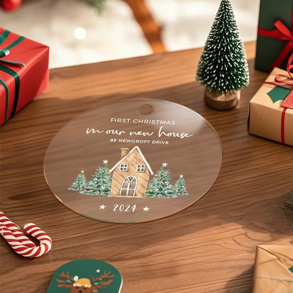 A front view of the personalized Christmas ornament featuring a house surrounded by Christmas trees with the text "First Christmas in our new home," laying flat on a table, ready to be customized with the address and year.