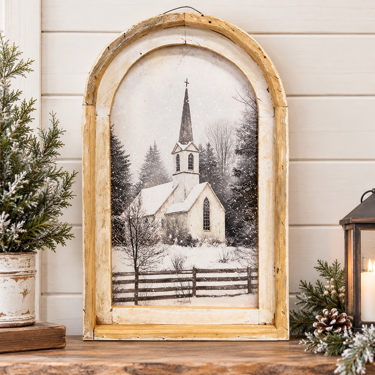 A rustic distressed wood arched frame wall decor featuring a black and white winter scene of a church on a snowy hill, styled on a wooden table with holiday greenery and a lantern.