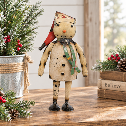A rustic handmade wooden snowman figurine wearing a red and green plaid Santa hat and a green striped scarf, standing on a wooden table surrounded by holiday greenery and books.