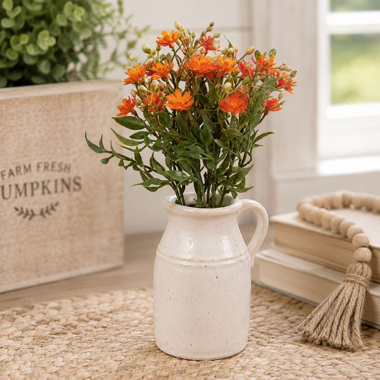 A small ceramic pitcher vase filled with orange flowers and green stems, styled on a woven rug beside stacked books, a wooden bead garland, and a burlap box labeled 'Farm Fresh Pumpkins' in a bright indoor setting.