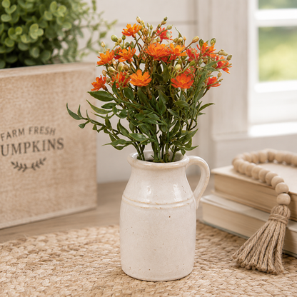 A small ceramic pitcher vase filled with orange flowers and green stems, styled on a woven rug beside stacked books, a wooden bead garland, and a burlap box labeled 'Farm Fresh Pumpkins' in a bright indoor setting.