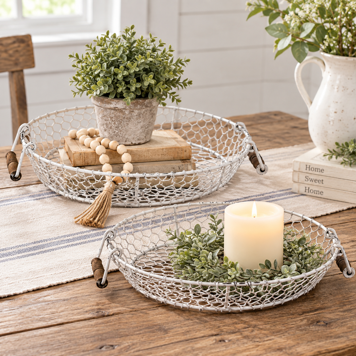 Two round white wire serving trays with dark wooden handles are styled on a rustic wooden table. One tray holds a small potted green plant, a wooden bead garland, and books, while the other holds a lit cream pillar candle in a green leafy wreath.