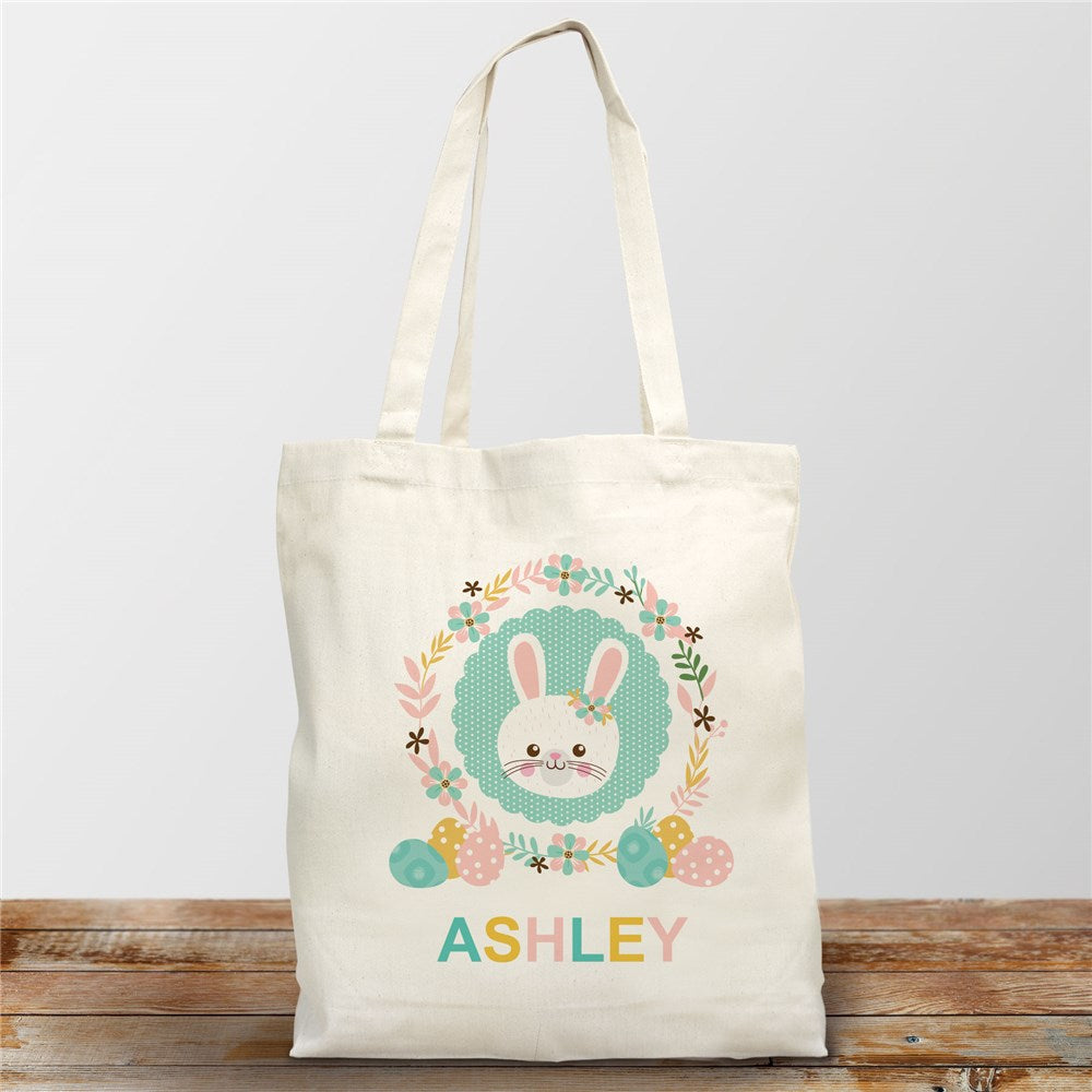 Personalized Easter Bunny Wreath Tote Bag on display – A cotton canvas tote bag featuring a printed bunny surrounded by a floral and egg wreath in pastel pink, marigold, and teal blue, with a custom embroidered name.

Easter tote bag ready for an egg hunt – A soft off-white canvas tote with a festive bunny wreath design, reinforced straps, and space for personalized embroidery, perfect for collecting eggs and Easter treats.