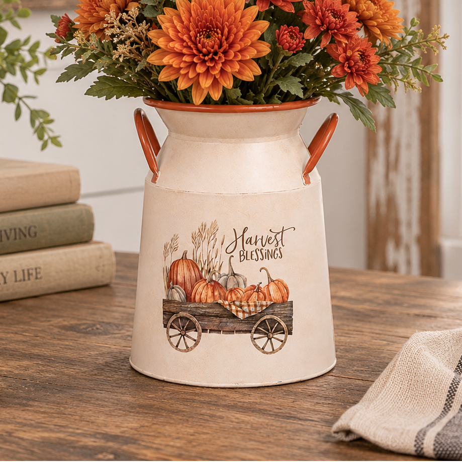 A cream-colored decorative milk canister planter with orange handles and a fall harvest illustration, filled with orange and red chrysanthemums, displayed on a wooden table in a rustic home setting.