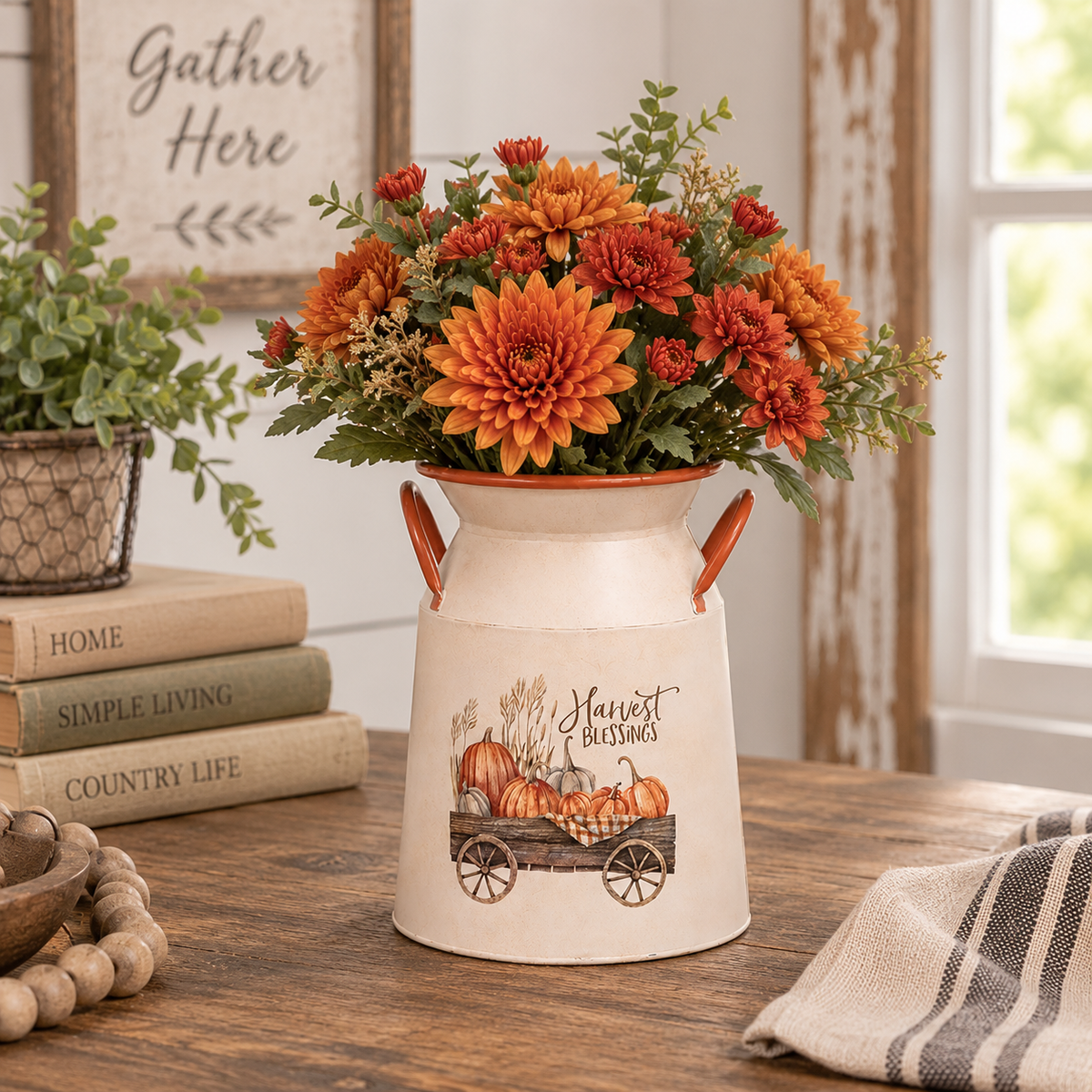 A cream-colored decorative milk canister planter with orange handles and a fall harvest illustration, filled with orange and red chrysanthemums, displayed on a wooden table in a rustic home setting.