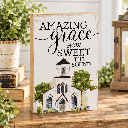 A rectangular decorative sign with a distressed white background stands on a wooden table. The sign reads “AMAZING grace HOW SWEET THE SOUND” in black script and serif lettering above a painted church with green trees and small purple flowers.