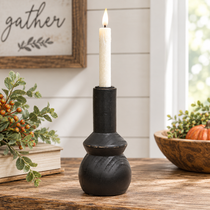 A black rustic ceramic candle holder with a lit white pillar candle, displayed on a wooden table in a cozy home setting with fall decorations.