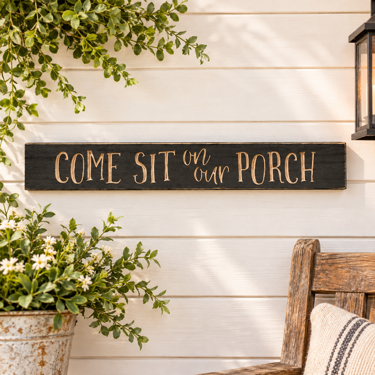 A rustic black wooden sign with the phrase 'COME SIT on our PORCH' in a warm beige script, mounted on a white wood-paneled wall. The scene is styled with a potted plant in the lower left, a wooden bench in the lower right, and a hanging lantern in the upper right.