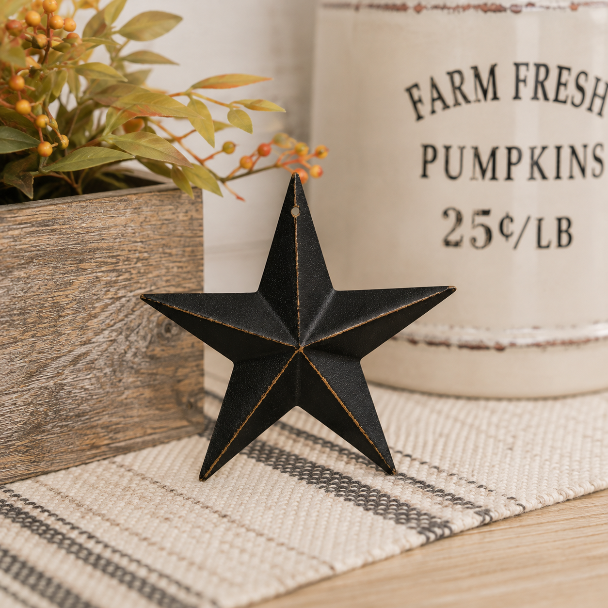 A black metal five-point star ornament with gold-toned edge detailing, displayed on a woven beige runner in a rustic setting with a wooden planter and a white ceramic mug in the background.