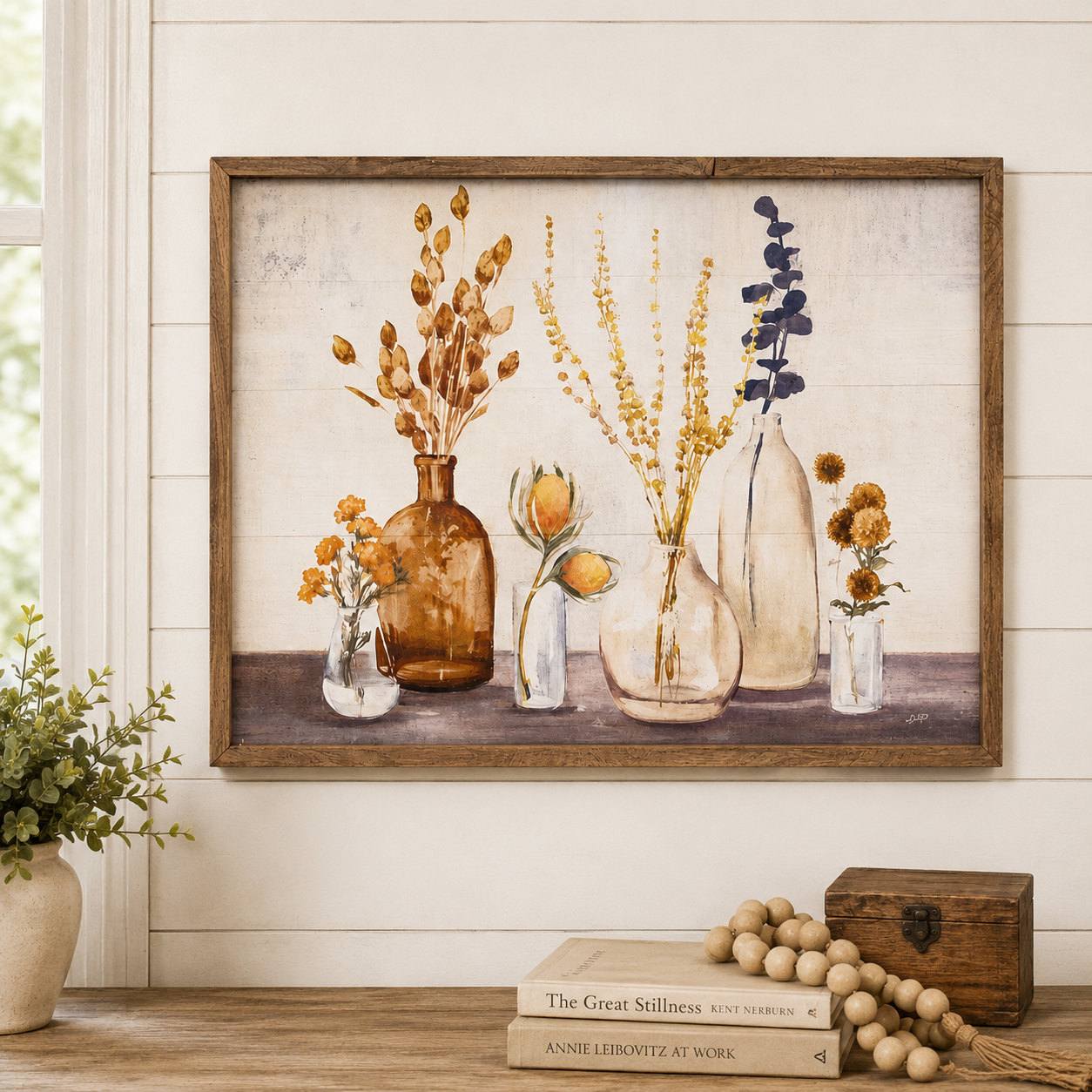 A framed rustic art print featuring a still life of dried flowers and botanicals in glass vases, displayed on a wooden shelf against a white shiplap wall with decorative books, a small wooden box, and a potted plant.