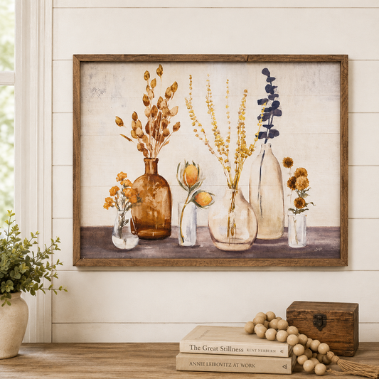 A framed rustic art print featuring a still life of dried flowers and botanicals in glass vases, displayed on a wooden shelf against a white shiplap wall with decorative books, a small wooden box, and a potted plant.