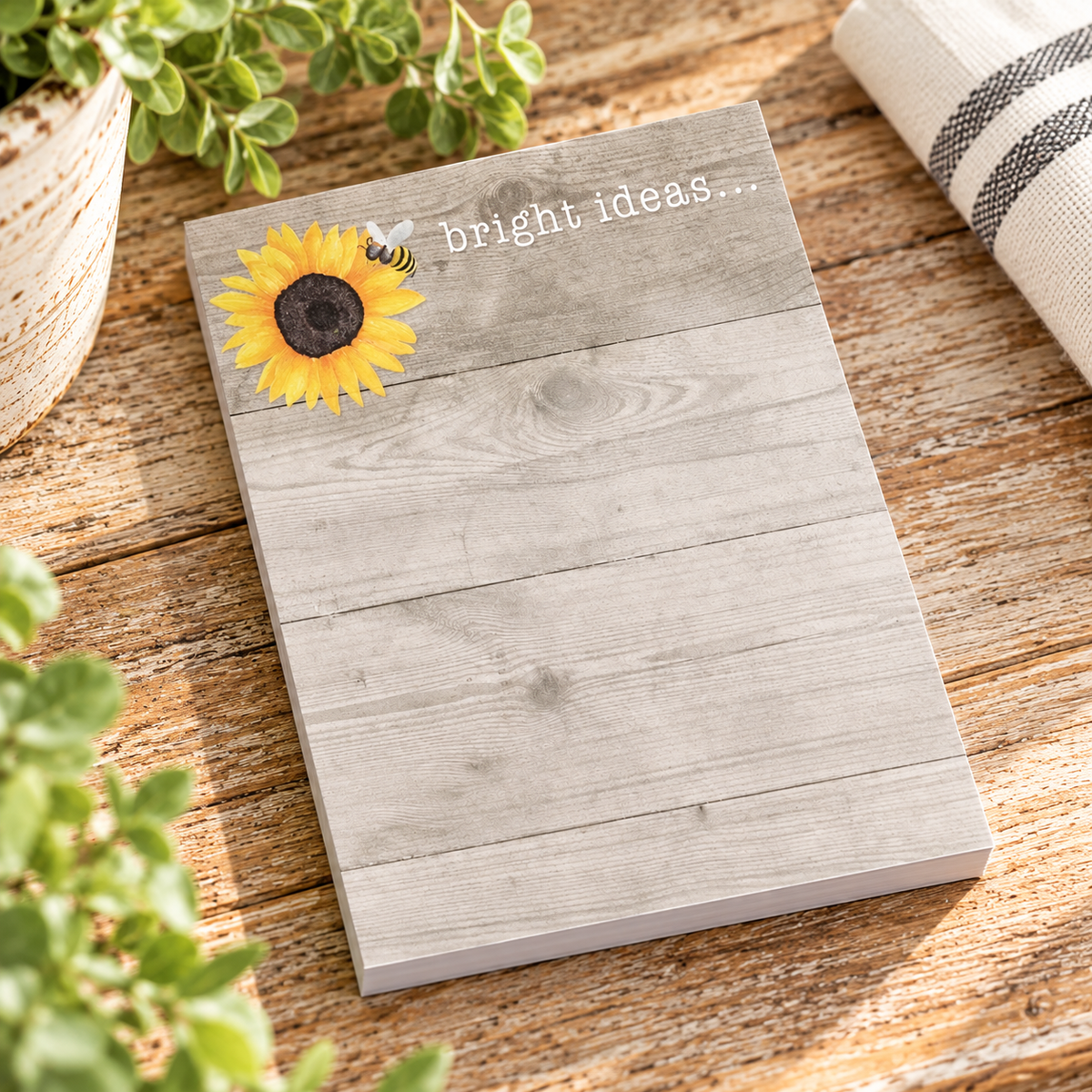 A rectangular notepad with a rustic gray wood plank design and a yellow sunflower with a bee near the top left, placed on a wooden surface with plants and a striped cloth around it.