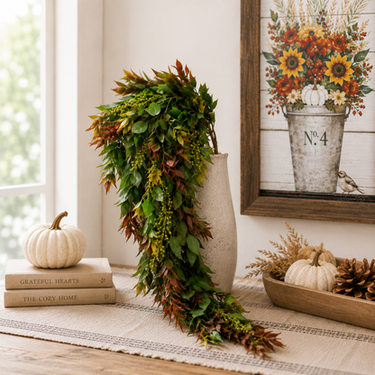 An autumn-themed faux leaf garland in shades of green, brown, and orange, draped over a beige ceramic vase on a rustic table runner. The scene is styled with a small white pumpkin on stacked books, a wooden tray with pinecones and mini pumpkins, and a framed floral print in the background.