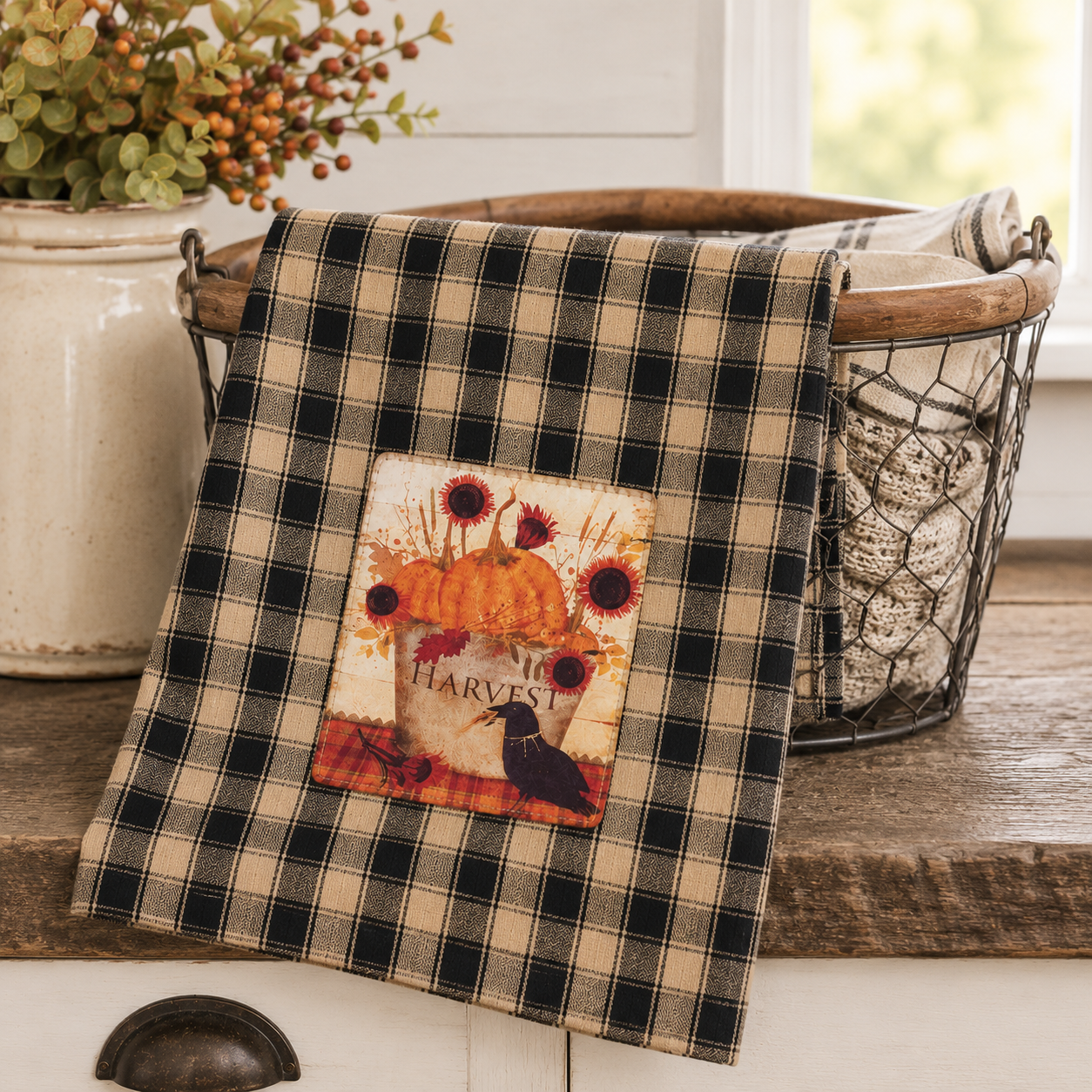 A black and beige plaid kitchen towel draped over a rustic wire basket on a wooden counter, featuring a central autumn-themed patch with pumpkins, sunflowers, and a raven.