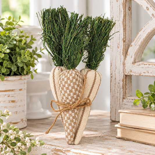 A decorative carrot-shaped fabric vase or planter with two green pine sprigs inserted at the top, tied with twine, displayed on a rustic wooden surface with potted plants and books in the background.