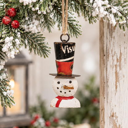 A small rustic Christmas ornament shaped like a snowman hanging from a tree branch. The ornament has a black top hat with red and white painted sections, a white snowman body, and a red scarf.