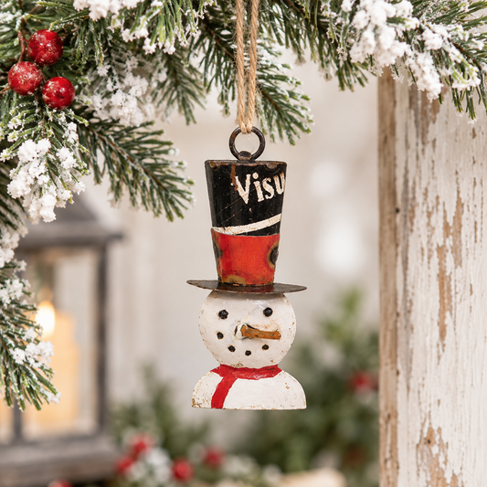 A small rustic Christmas ornament shaped like a snowman hanging from a tree branch. The ornament has a black top hat with red and white painted sections, a white snowman body, and a red scarf.