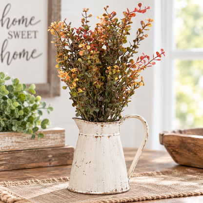 A distressed white metal pitcher vase filled with artificial autumn foliage, styled on a burlap runner on a wooden table with rustic home decor in the background.