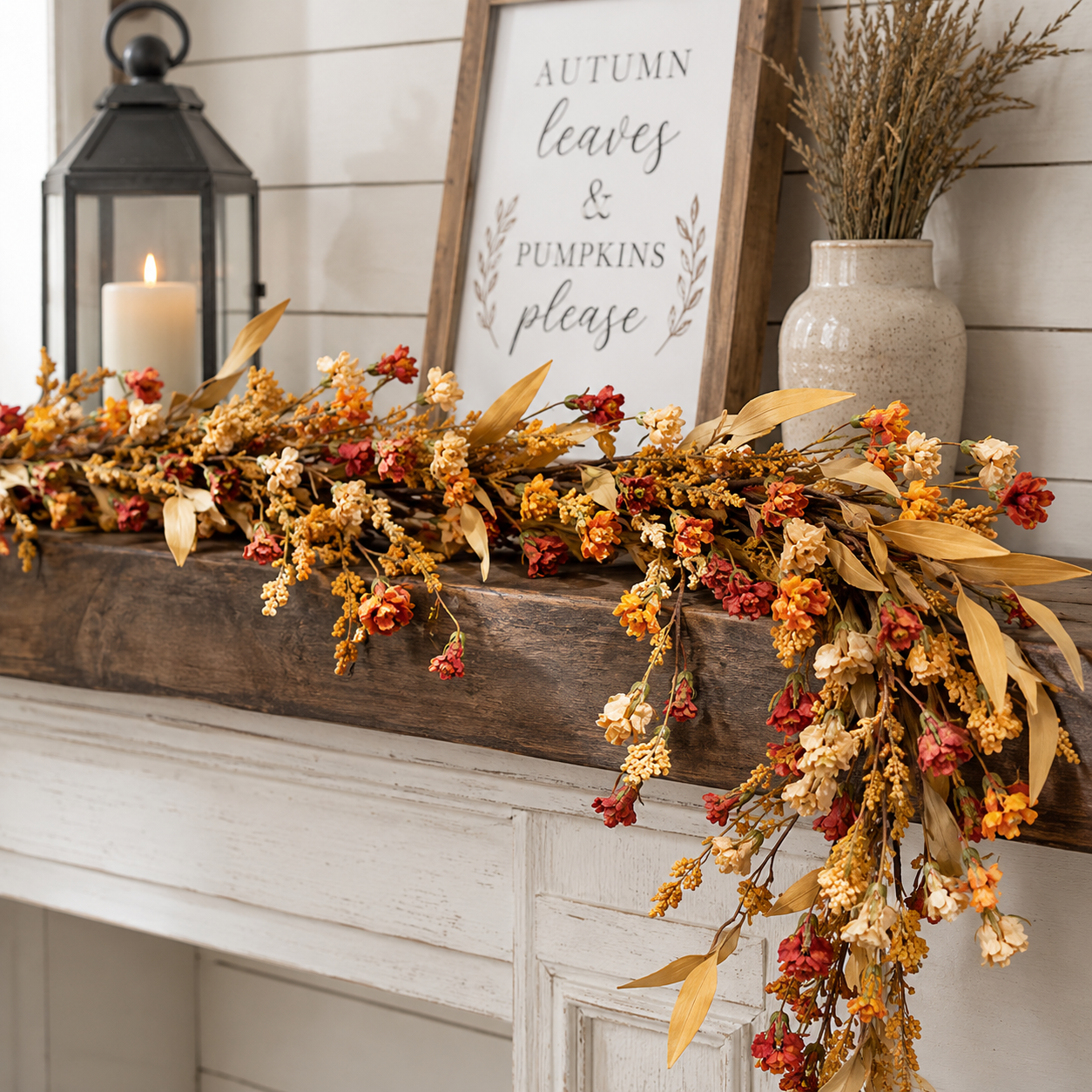 An autumn floral garland with dried leaves, berries, and small flowers draped across a rustic wooden mantel in a styled home setting. A framed sign reading 'Autumn leaves & pumpkins please' hangs on the wall, with a candle lantern and a vase of dried grass nearby.
