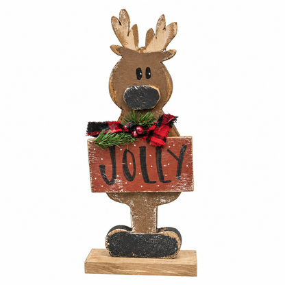 A rustic wooden Christmas reindeer decoration holding a sign that says 'JOLLY' in black letters on a distressed reddish-brown background. The reindeer has a natural brown wood finish with black painted details, and is decorated with a red and black plaid scarf, green pine branches, and small red berries.