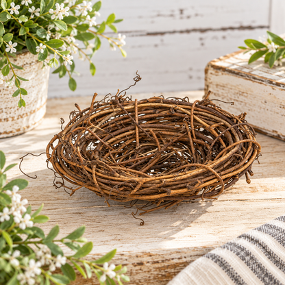 A decorative twig nest bowl made of intertwined brown branches, placed on a light wood surface with floral props and a rustic book in the background.