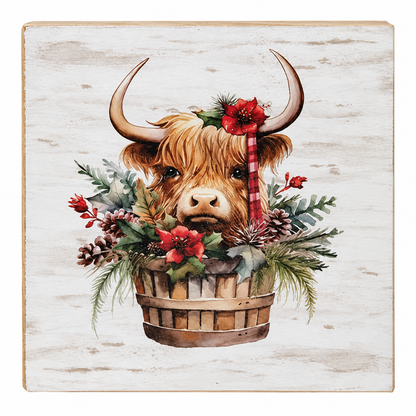 A square decorative wall plaque featuring a painted Highland cow head with curved horns, surrounded by a rustic wooden basket filled with red flowers, green foliage, pinecones, and a red-and-white striped candy cane, set against a distressed off-white wood background.