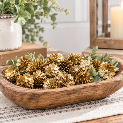 A wooden bowl filled with decorative gold pinecones and green eucalyptus leaves, styled on a table runner with a rustic candle and planter in the background.