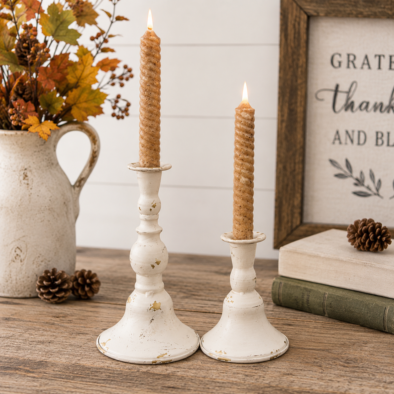 Two white distressed metal candlestick holders with lit taper candles, styled on a wooden tabletop with a ceramic pitcher of autumn leaves, pinecones, books, and a framed sign in the background.
