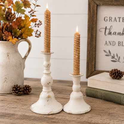 Two white distressed metal candlestick holders with lit taper candles, styled on a wooden tabletop with a ceramic pitcher of autumn leaves, pinecones, books, and a framed sign in the background.
