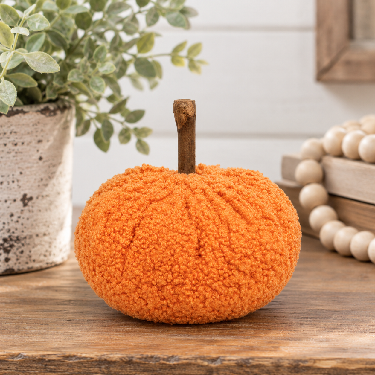 An orange textured yarn pumpkin decoration with a short brown stem, displayed on a wooden surface with a potted plant and wooden beads in the background.