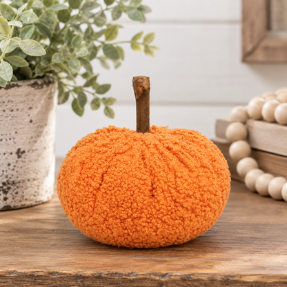 An orange textured yarn pumpkin decoration with a short brown stem, displayed on a wooden surface with a potted plant and wooden beads in the background.