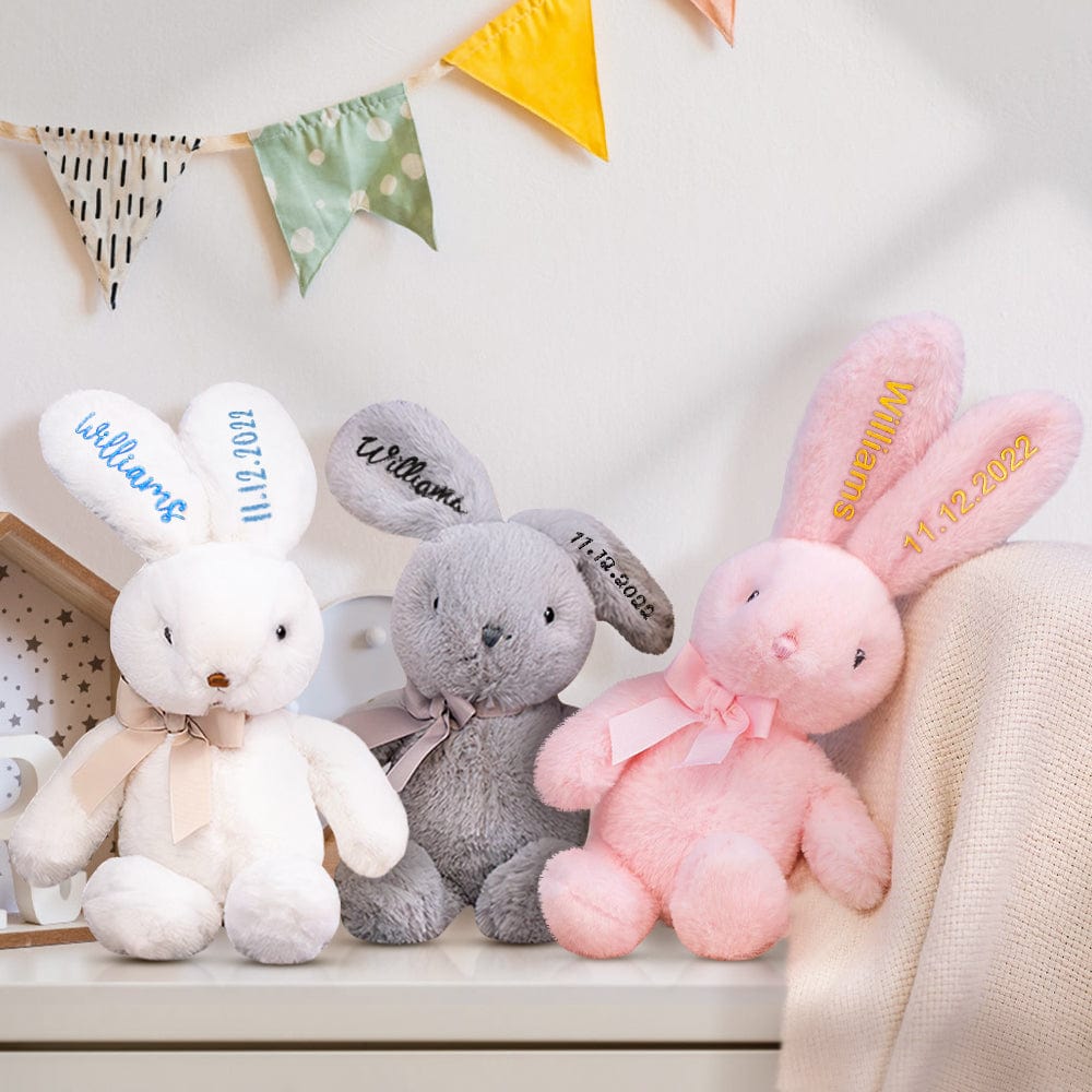 Three embroidered bunnies in a row – A white bunny with blue embroidery, a gray bunny with black embroidery, and a yellow bunny with yellow embroidery sitting together.