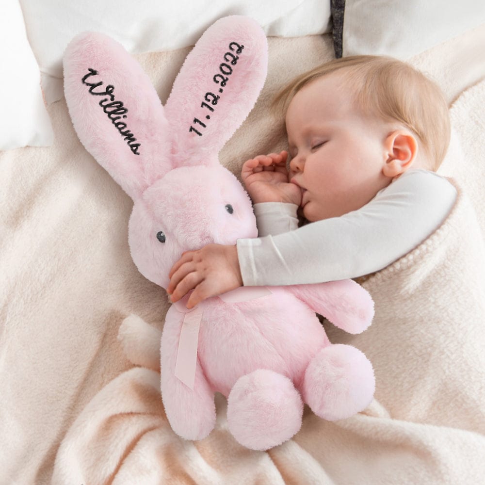 Sleeping baby holding a plush bunny – A baby cuddling a pink bunny with black embroidered details.