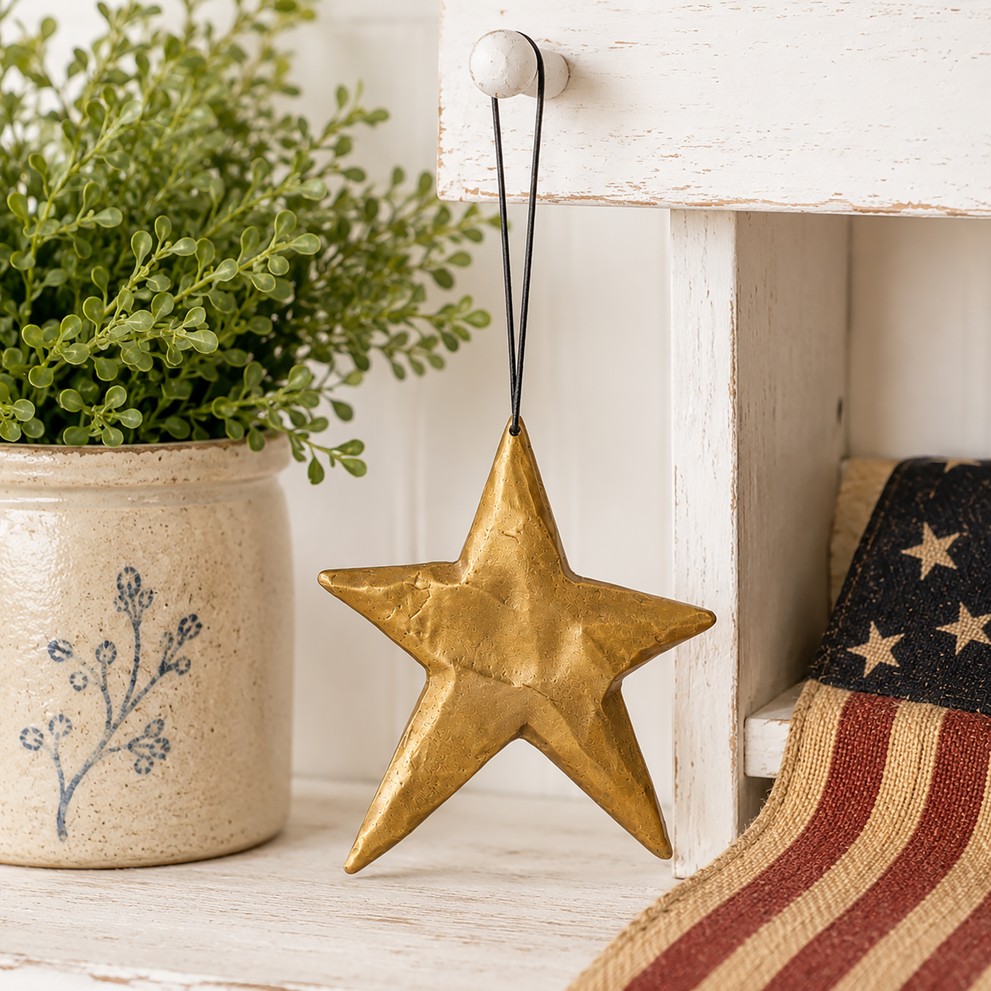 Antique gold star ornament hanging from a black cord on a white farmhouse peg beside greenery and primitive Americana décor in a bright rustic setting.