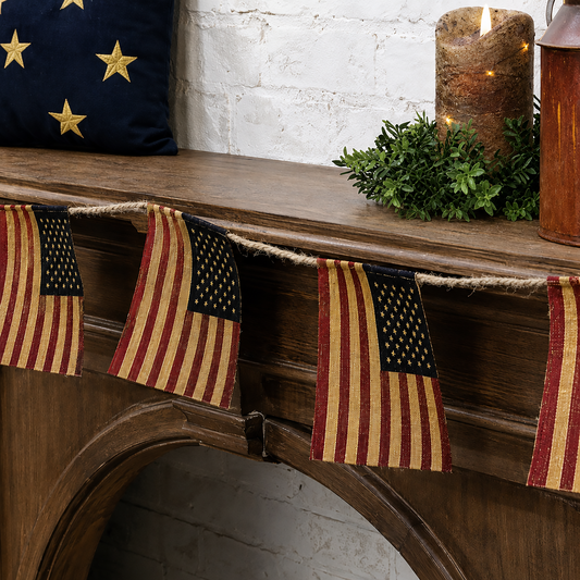 Vintage-inspired American flag garland made of muted red, navy, and beige burlap flags hanging from a jute rope across a rustic wooden mantel with patriotic farmhouse décor.