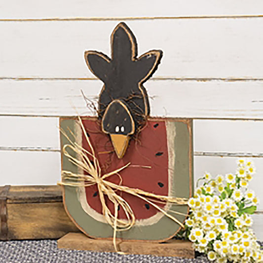 Wooden watermelon slice with crow on top, rustic painted finish in red, green, and black, with raffia and twig accents for primitive summer decor